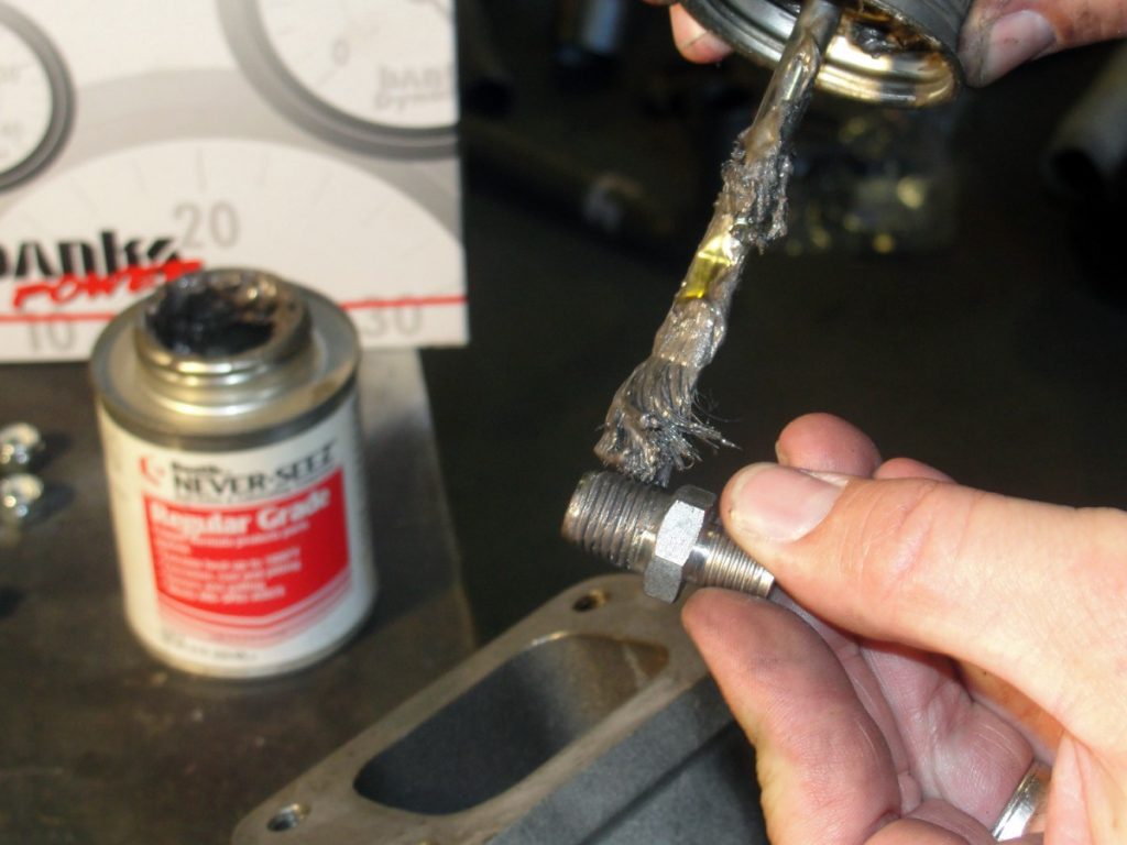 A pyrometer probe adapter is installed in the new exhaust manifold using anti-seize on the threads. The new exhaust manifold can then be installed using the original mounting bolts. The Banks instructions recommend that the bolts should be inserted into the manifold and then secured using rubber bands until the bolts are started. We thought we knew a better way and didn’t need the rubber bands. After several failed attempts due to bolts falling out and a good bit of cursing, we can say from experience that a small package of rubber bands is money well spent! Once the manifold is in place the glow plugs can be re-installed and the glow plug wiring and fuel return lines between the injectors should be routed up and away from the manifold. (photo ref: rubber cement)